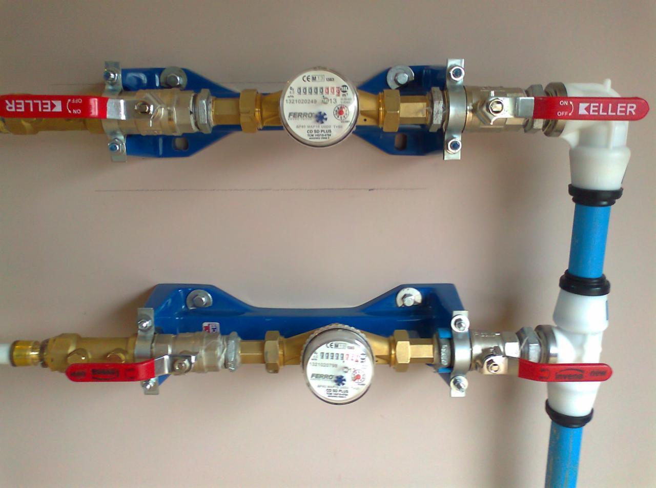 Dwa wodomierze FERRO, z niebieskimi uchwytami, zamontowane na ścianie. Instalacja z rur Keller z czerwonymi zaworami. Widoczne połączenia i śruby.