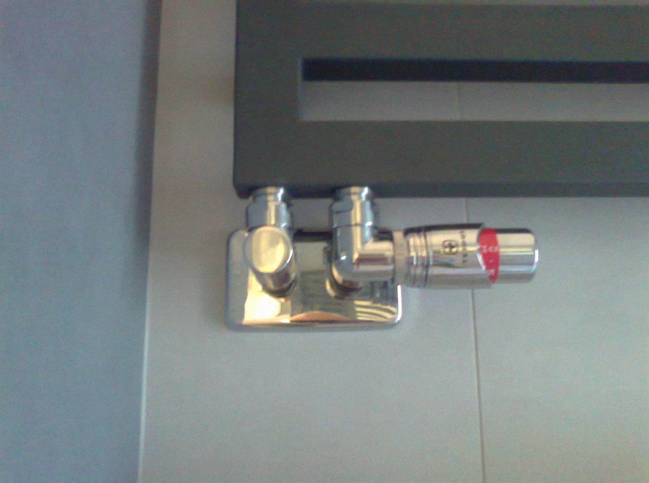 Błyszczący, chromowany zawór termostatyczny przy grzejniku drabinkowym, zamontowany na jasnych płytkach. Widoczne detale instalacji grzewczej i regulacja temperatury.