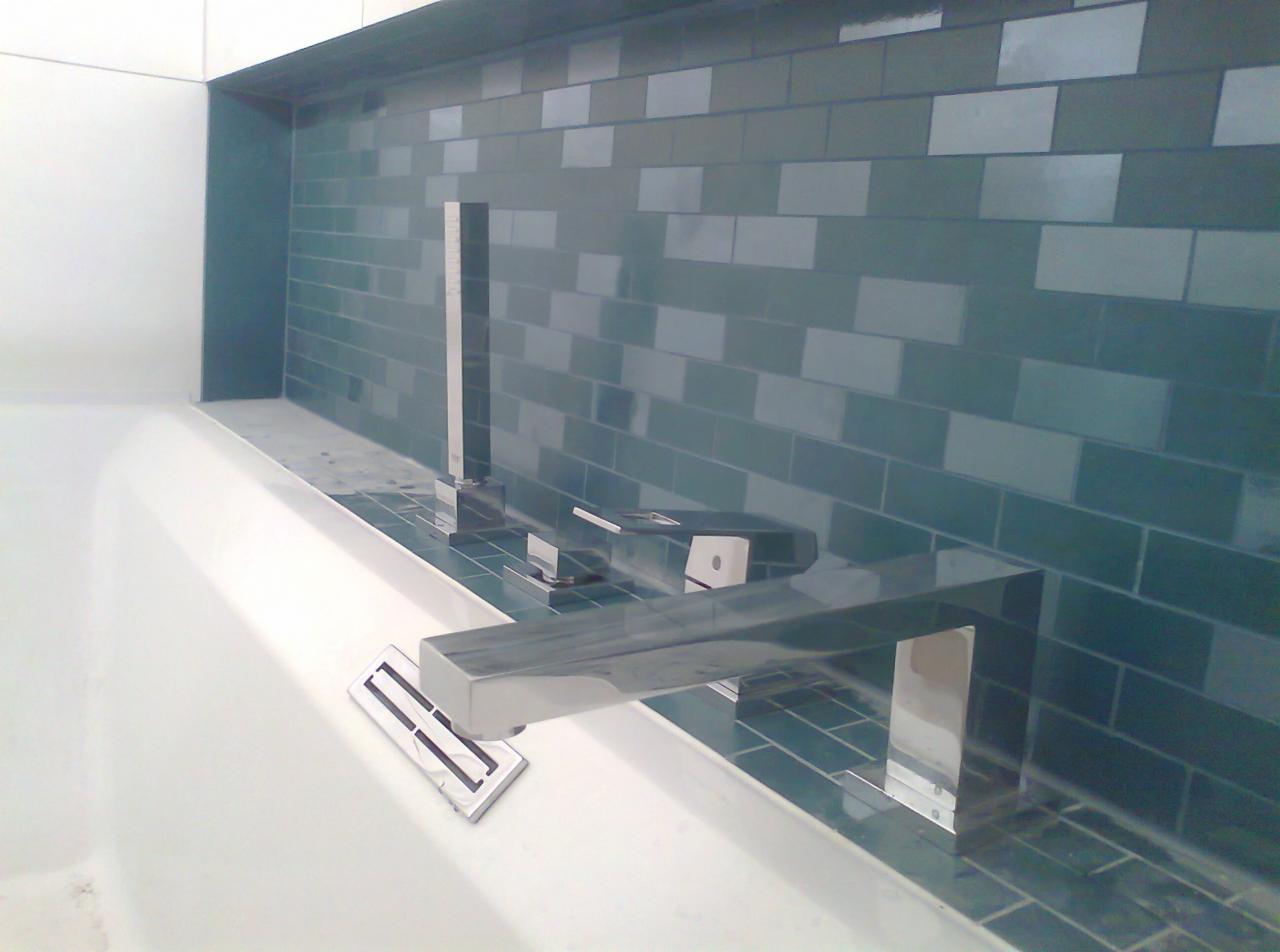 Bateria wannowa chromowana, zamontowana na tle ściany wyłożonej turkusową, prostokątną mozaiką. Widoczny fragment białej wanny z odpływem liniowym.