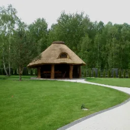 Kryta strzechą konstrukcja ogrodowa z drewnianymi filarami, otoczona zielonym trawnikiem, drzewami i ścieżką z kostki brukowej.