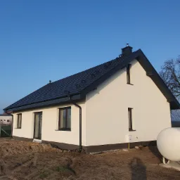 Nowoczesny dom jednorodzinny z ciemną dachówką i białymi ścianami, widoczny zbiornik na gaz ziemny z logo GASPOL na tle błękitnego nieba.