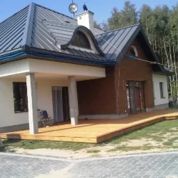 Nowoczesny dom jednorodzinny z drewnianym tarasem, widok z zewnątrz na elewację z drewnianą okładziną i szarym dachem.