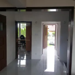 Wnętrze domu po remoncie: widok na kilka otworów drzwiowych z zamontowanymi ościeżnicami i drzwiami w kolorze ciemnego brązu, z błyszczącą podłogą z płytek, w tle widoczne inne pomieszczenia...