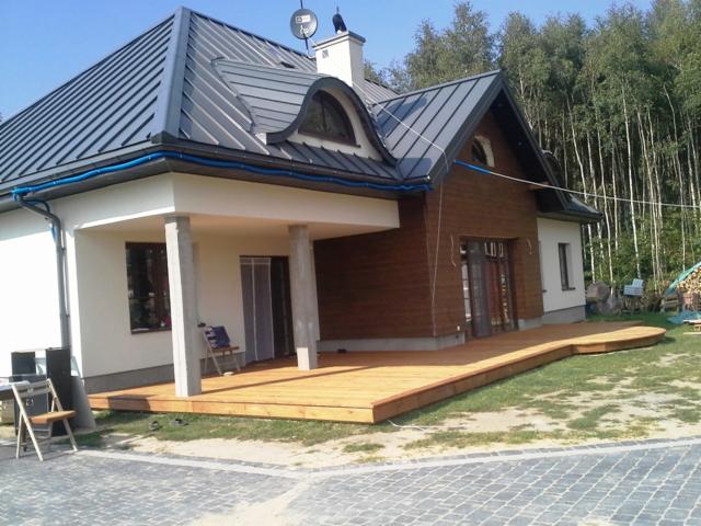 Nowoczesny dom jednorodzinny z drewnianym tarasem, widok z zewnątrz na elewację z drewnianą okładziną i szarym dachem.