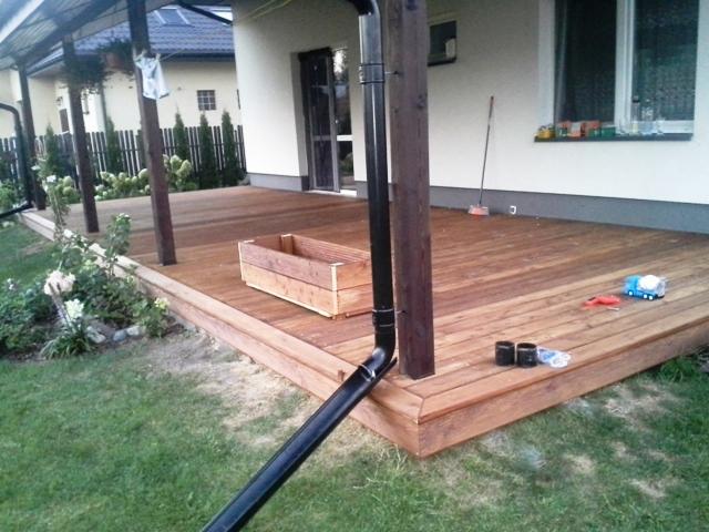 Drewniany taras przydomowy z zadaszeniem, widok z poziomu trawnika, z widoczną rynną, drewnianą skrzynką na kwiaty i zabawkami dziecka.