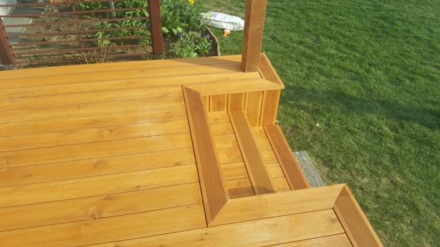 Drewniany taras z widocznymi schodami, wykończony w ciepłym odcieniu brązu, otoczony zielonym trawnikiem i fragmentem kwietnika.