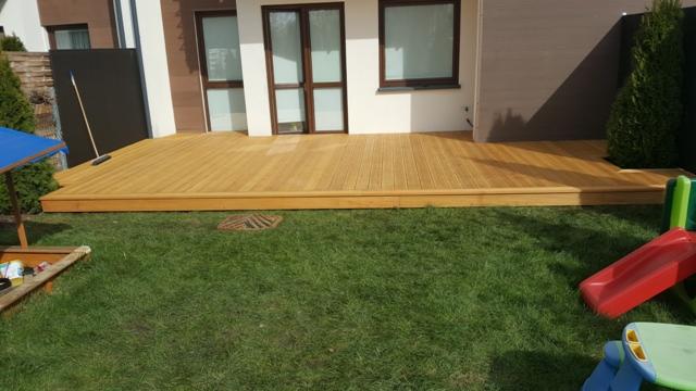 Wykończony taras z drewna na poziomie gruntu, przylegający do domu z widocznymi drzwiami balkonowymi i oknami, z elementami placu zabaw dla dzieci na pierwszym planie.