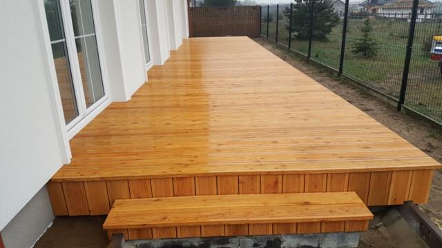 Długi, drewniany taras przylegający do białego budynku z oknami, częściowo mokry od deszczu, z niskim stopniem wejściowym i ogrodzeniem w tle.