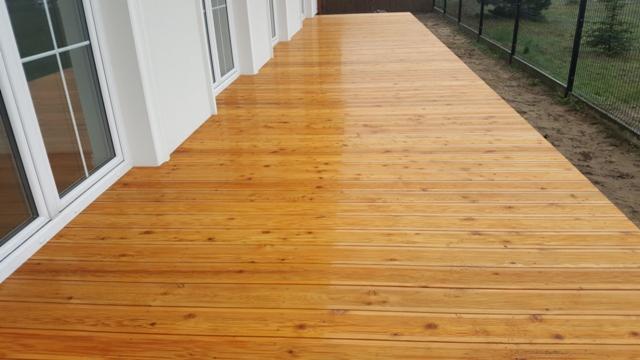 Świeżo polakierowany drewniany taras przy białej ścianie budynku z widocznymi oknami, część tarasu jeszcze mokra od lakieru, w tle ogrodzenie z siatki.