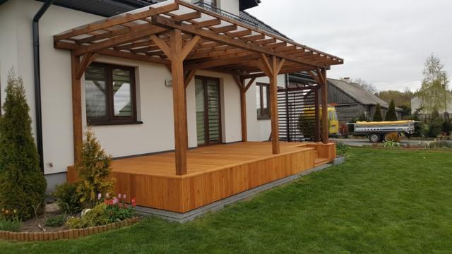 Drewniana konstrukcja zadaszonej werandy przylegająca do białego domu z oknami, obok zadbany trawnik z rabatą kwiatową, w tle zabudowania gospodarcze i przyczepa.