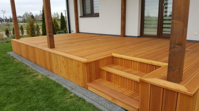 Drewniany taras z jasnego drewna z niskimi schodkami, otoczony zielonym trawnikiem, przylegający do białego budynku z oknem z roletami.