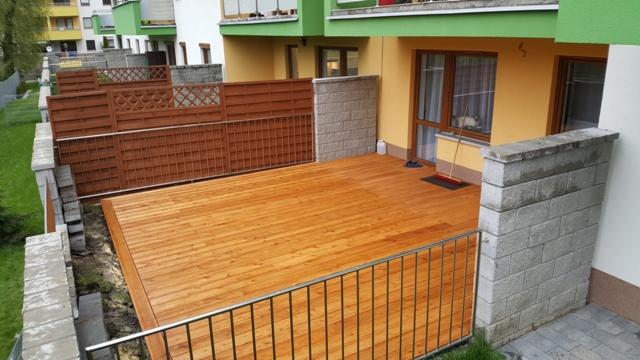 Jasna, drewniana podłoga tarasu z metalową barierką, otoczona niskim murem z bloczków betonowych i panelami z brązowego drewna, przylegająca do budynku mieszkalnego z zielonymi i żółtymi elementami...