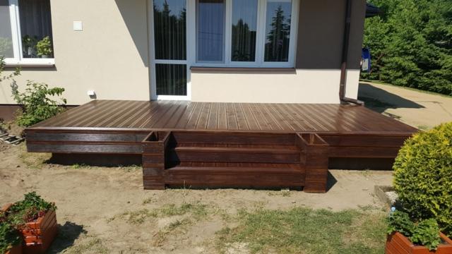 Wykonany z ciemnego drewna taras przydomowy z wbudowanymi donicami, widok z poziomu gruntu, na tle okna tarasowego.