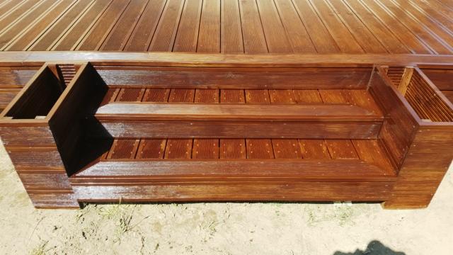 Drewniana konstrukcja schodów z dwoma stopniami i wbudowanymi donicami po bokach, wykonana z ciemnobrązowego drewna z widoczną strukturą słojów, ułożona na piasku.