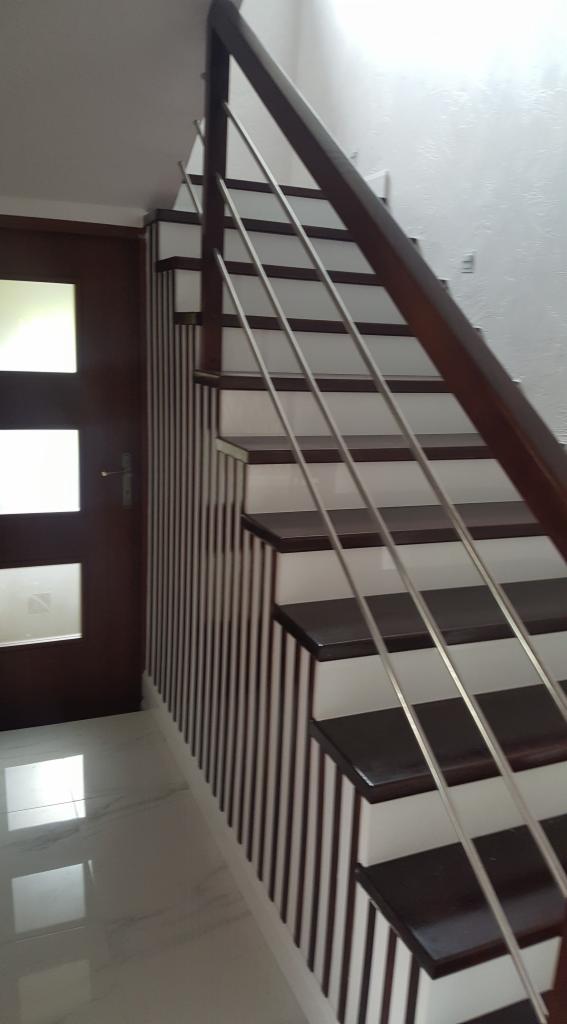 Nowoczesne schody drewniane z metalową balustradą, oświetlone naturalnym światłem, z widoczną częścią drzwi z przeszkleniami i błyszczącą podłogą.