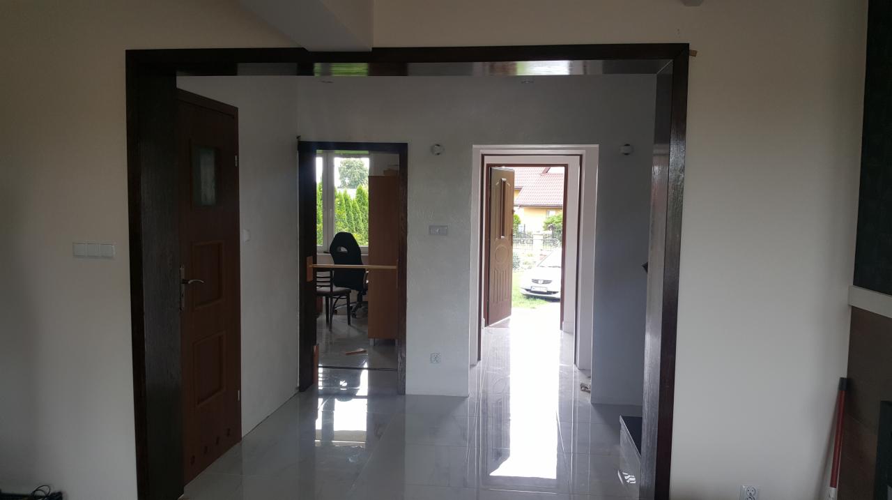 Wnętrze domu po remoncie: widok na kilka otworów drzwiowych z zamontowanymi ościeżnicami i drzwiami w kolorze ciemnego brązu, z błyszczącą podłogą z płytek, w tle widoczne inne pomieszczenia...