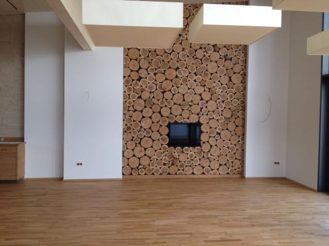 Ściana wewnętrzna wykończona okrągłymi kawałkami drewna z wkomponowanym czarnym prostokątnym wkładem kominkowym, drewniana podłoga, białe ściany i podwieszany sufit.