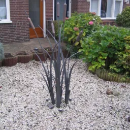 Metalowa dekoracja ogrodowa w kształcie pałki wodnej na tle domu z czerwonej cegły i żwirowego podłoża z oświetleniem punktowym.