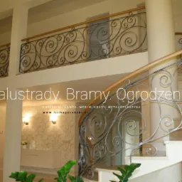 Wnętrze domu z balustradą schodową i balustradą na antresoli, wykonaną z metalu o dekoracyjnych, spiralnych wzorach, z drewnianymi poręczami, widok z dołu schodów.