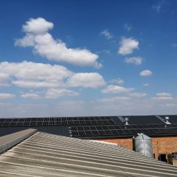 Montaż Kolektorów Słonecznych i Instalacji CO Andrzej Żmuda - Instalacja paneli słonecznych na dachu budynku gospodarczego z czerwonej cegły, widok z dachu sąsiedniego budynku, błękitne niebo z chmurami w tle.