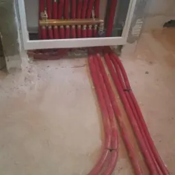 Rozdzielacz z czerwonymi rurami w ścianie podczas montażu systemu ogrzewania podłogowego, widoczne surowe wykończenie ściany i podłogi.