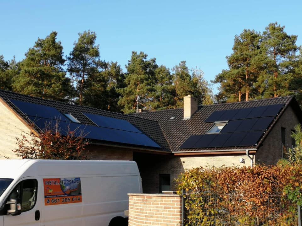Dom jednorodzinny z zamontowanymi panelami słonecznymi na dachu; widoczny biały samochód dostawczy z naklejką firmy instalacyjnej.