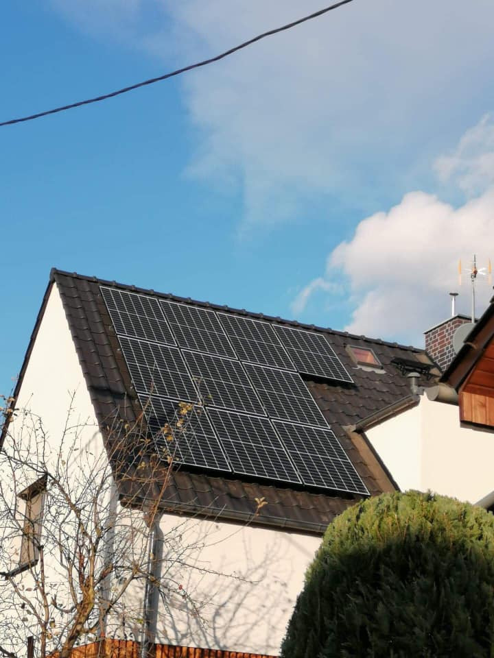 Panele fotowoltaiczne na dachu dwuspadowym białego domu jednorodzinnego, widok z dołu, błękitne niebo z chmurami w tle.
