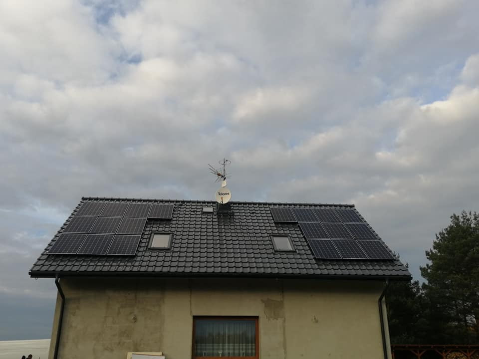 Dom z szarego tynku z panelami słonecznymi na czarnym dachu, widoczne okna dachowe i antena satelitarna Televes, pochmurne niebo w tle.