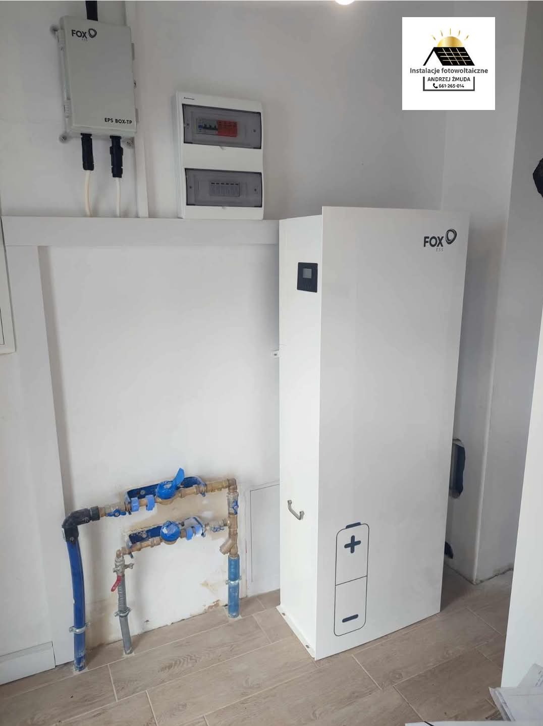 Biały akumulator energii z logo FOX ESS, zamontowany w pomieszczeniu z instalacją hydrauliczną i elektryczną, widoczny także rozdzielnica elektryczna i logo firmy fotowoltaicznej w rogu