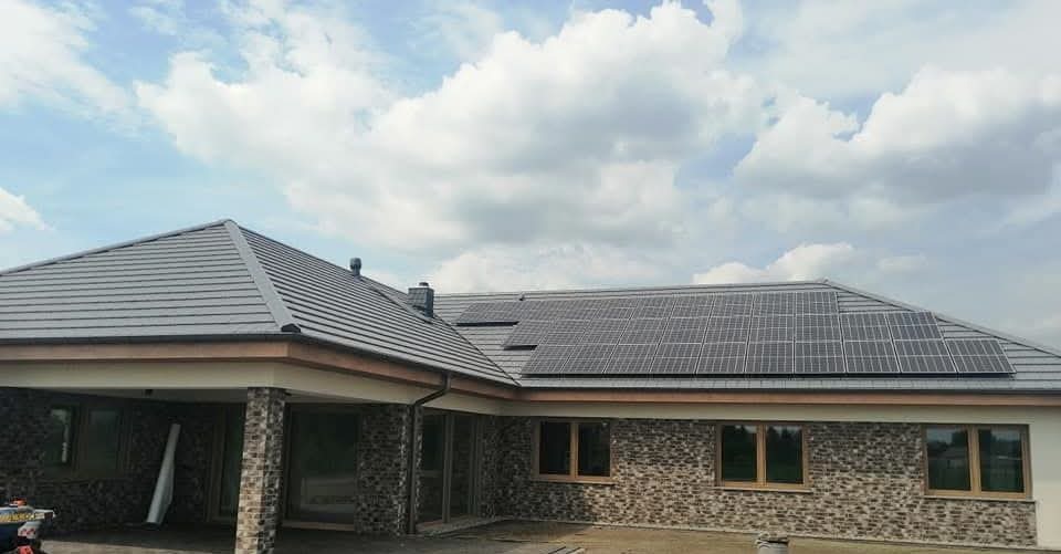 Dom jednorodzinny z szarą dachówką i panelami słonecznymi na dachu, widok z dołu na tle błękitnego nieba z chmurami.