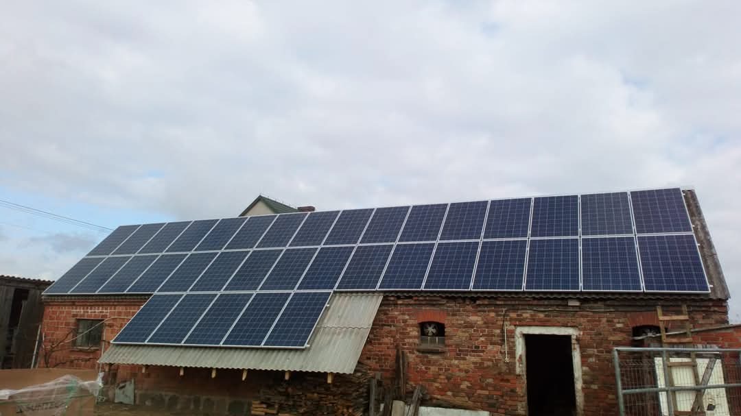 Panele słoneczne na dachu starego, ceglanego budynku gospodarczego z widocznymi śladami użytkowania i stertą drewna opałowego w pobliżu.