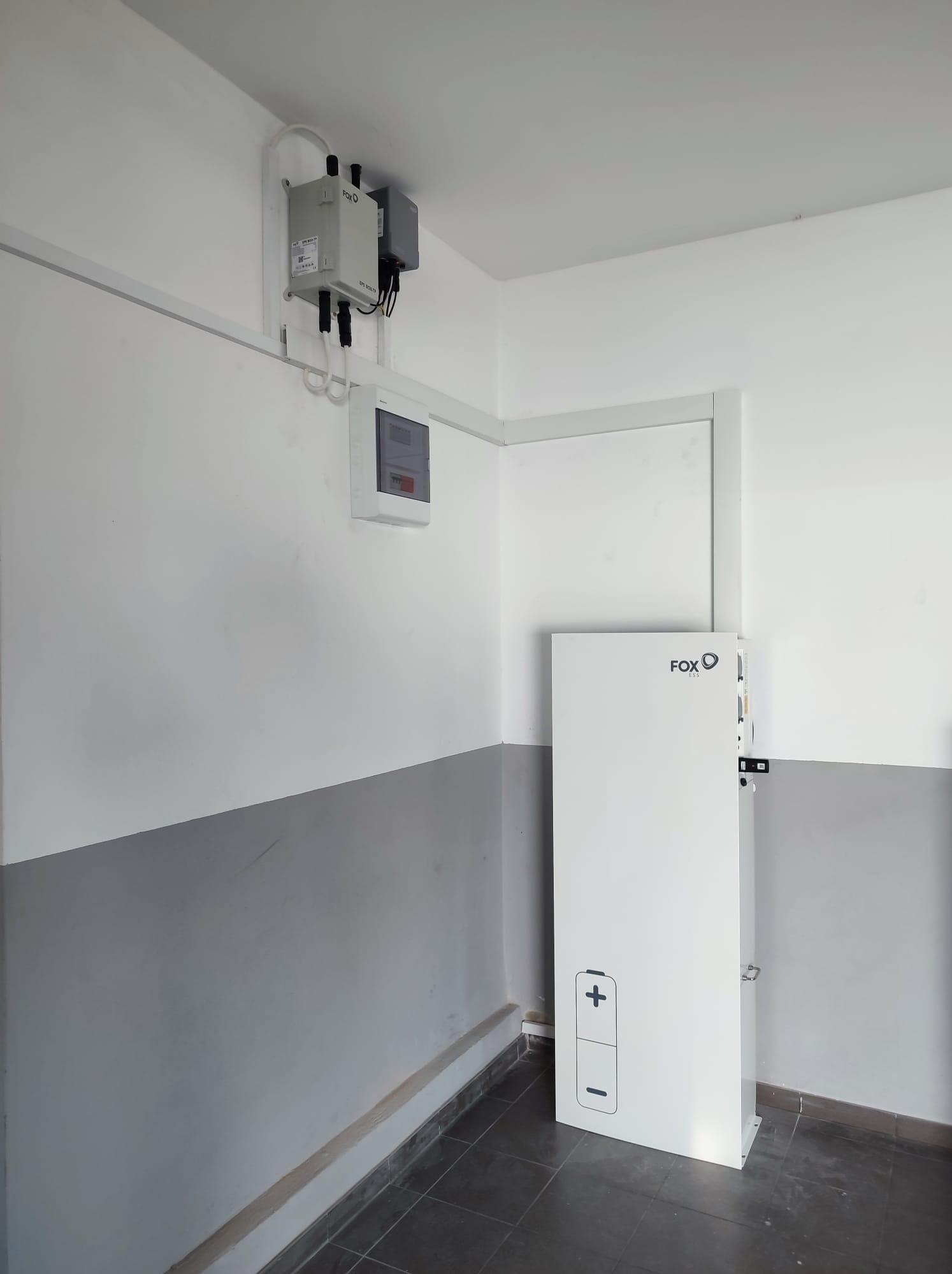 Biała bateria magazynu energii FoxESS z symbolem plus i minus, zamontowana w rogu pomieszczenia z szarą lamperią, obok skrzynka elektryczna i zabezpieczenia na ścianie.