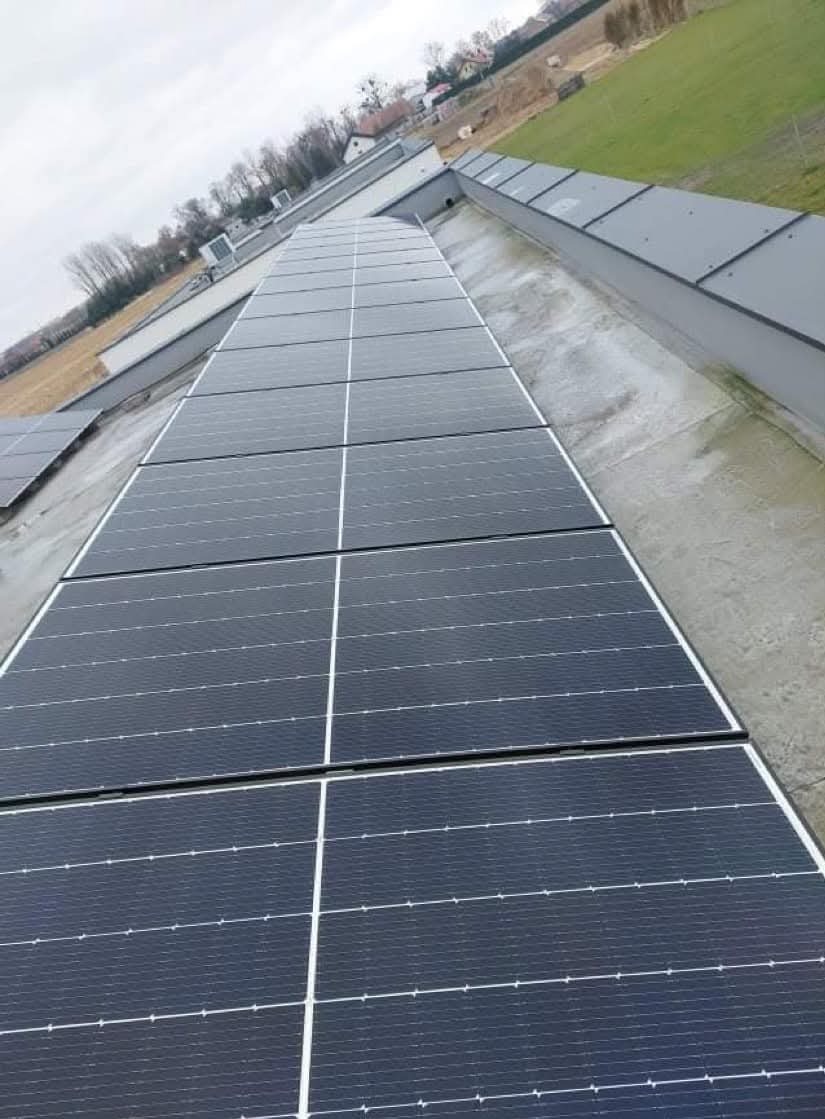 Rząd paneli fotowoltaicznych na płaskim dachu budynku komercyjnego, widok z góry, pochmurne niebo w tle.