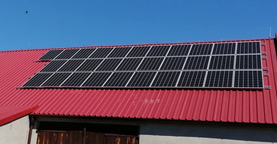 Panele słoneczne zainstalowane na falistej, czerwonej blasze dachu budynku gospodarczego w słoneczny dzień, widoczne zabezpieczenia przeciw ptakom.
