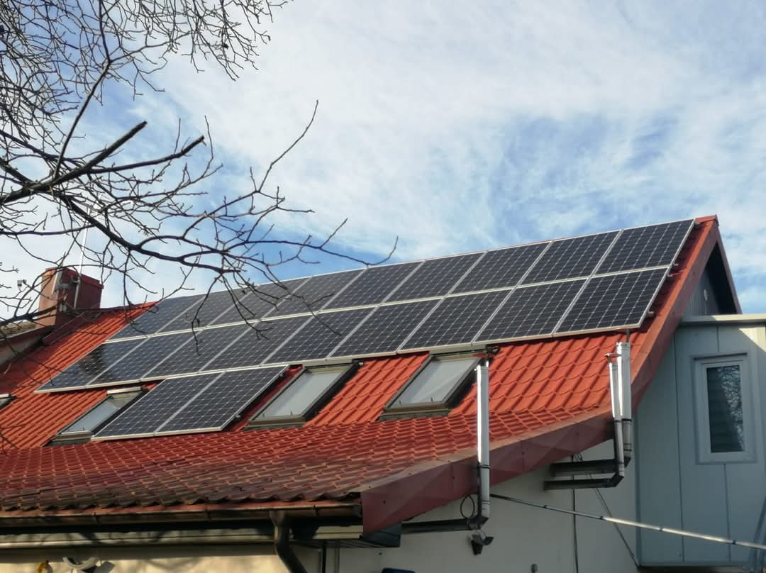 Panele słoneczne zamontowane na dachu z czerwonej dachówki, widoczne okna dachowe i fragment drzewa na tle błękitnego nieba z chmurami.