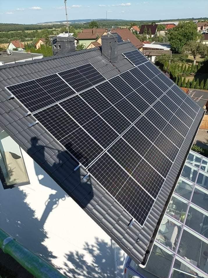 Panele słoneczne na dachu domu z ciemną dachówką, widoczny cień osoby instalującej oraz fragment ogrodu zimowego.