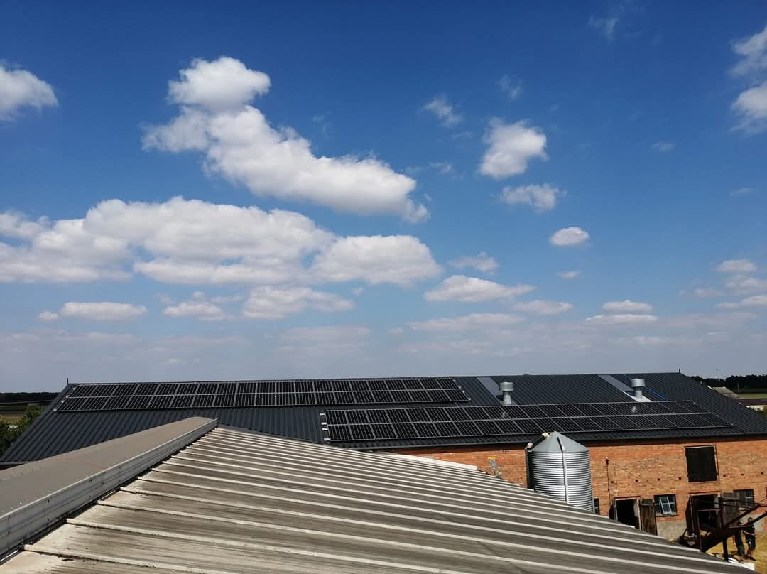 Instalacja paneli słonecznych na dachu budynku gospodarczego z czerwonej cegły, widok z dachu sąsiedniego budynku, błękitne niebo z chmurami w tle.