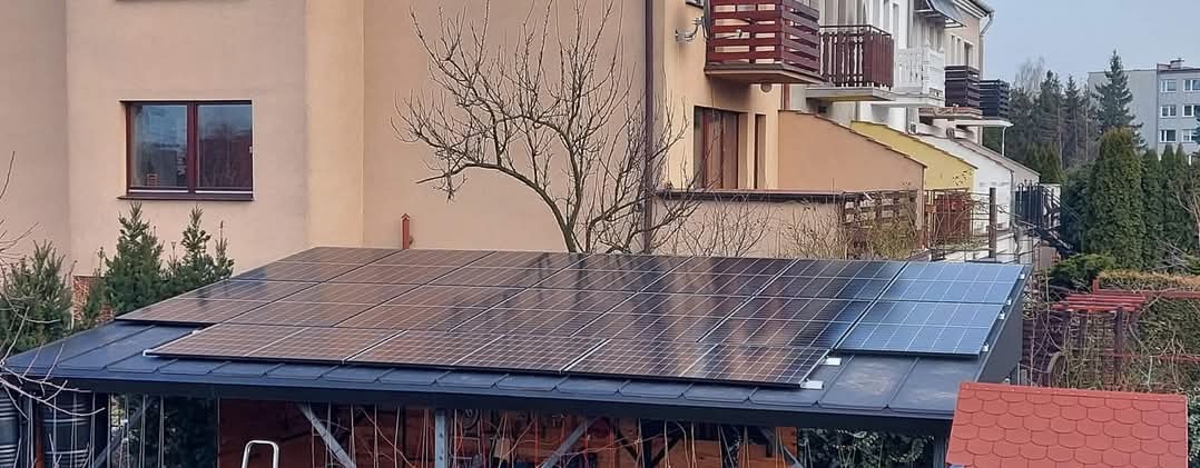 Panele słoneczne na dachu altany w ogrodzie, widoczne budynki mieszkalne w tle, pochmurne niebo.