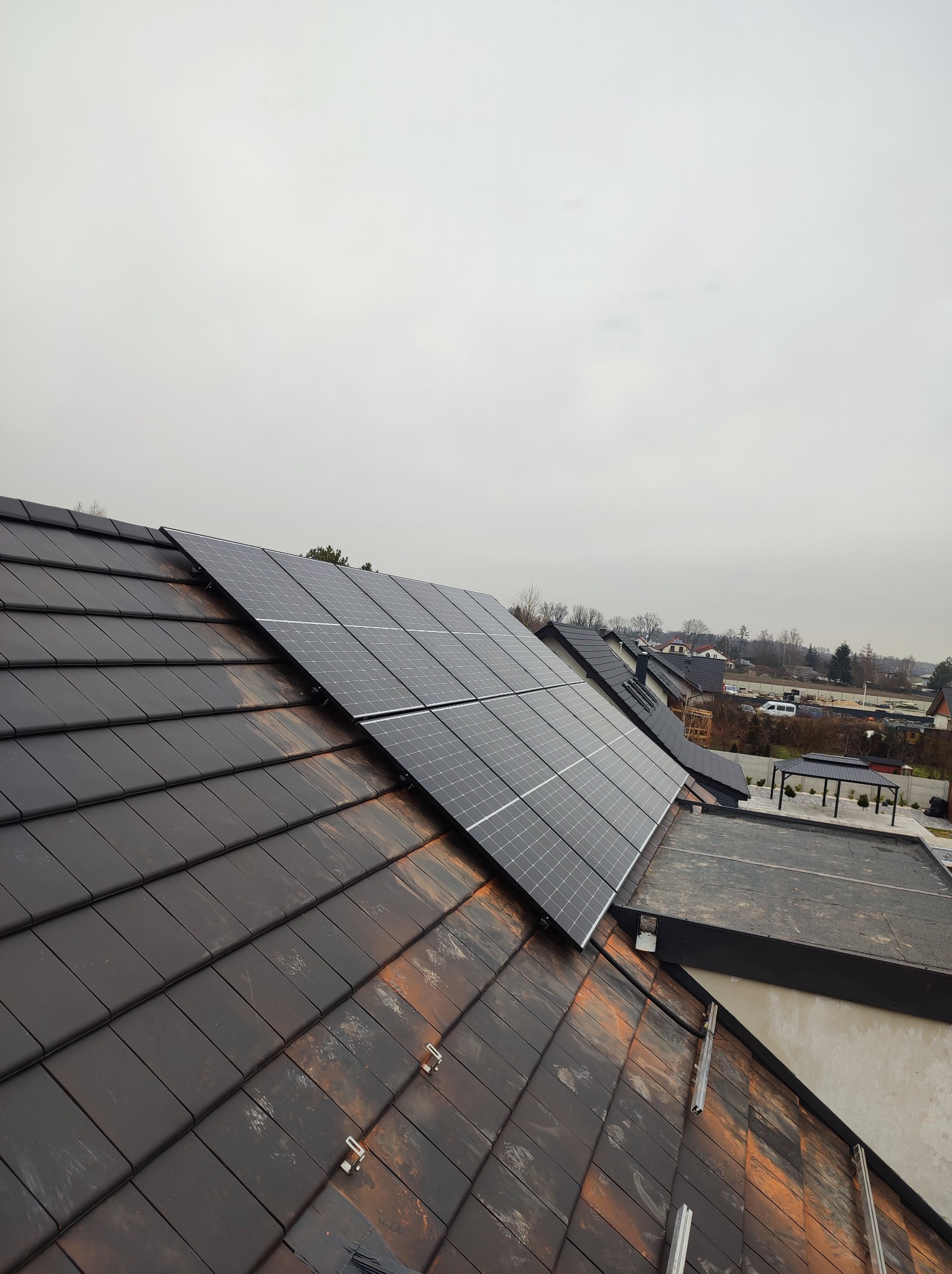 Montaż paneli fotowoltaicznych na dachu z brązowej dachówki ceramicznej, widoczne uchwyty montażowe, w tle zabudowa jednorodzinna i pochmurne niebo.