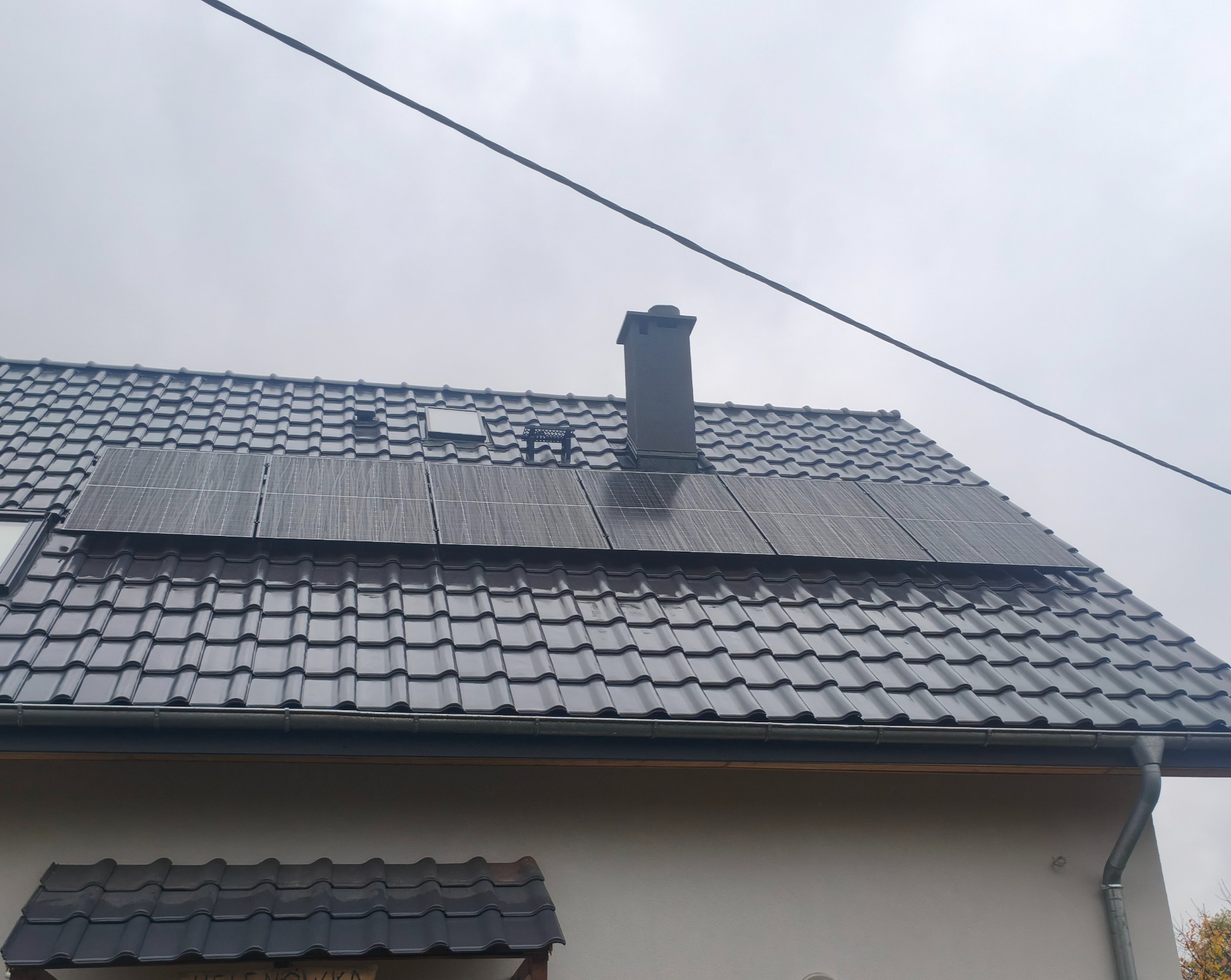 Ciemne panele słoneczne na grafitowym dachu domu jednorodzinnego z kominem, widoczne rynny i fragment elewacji. Pochmurne niebo w tle.