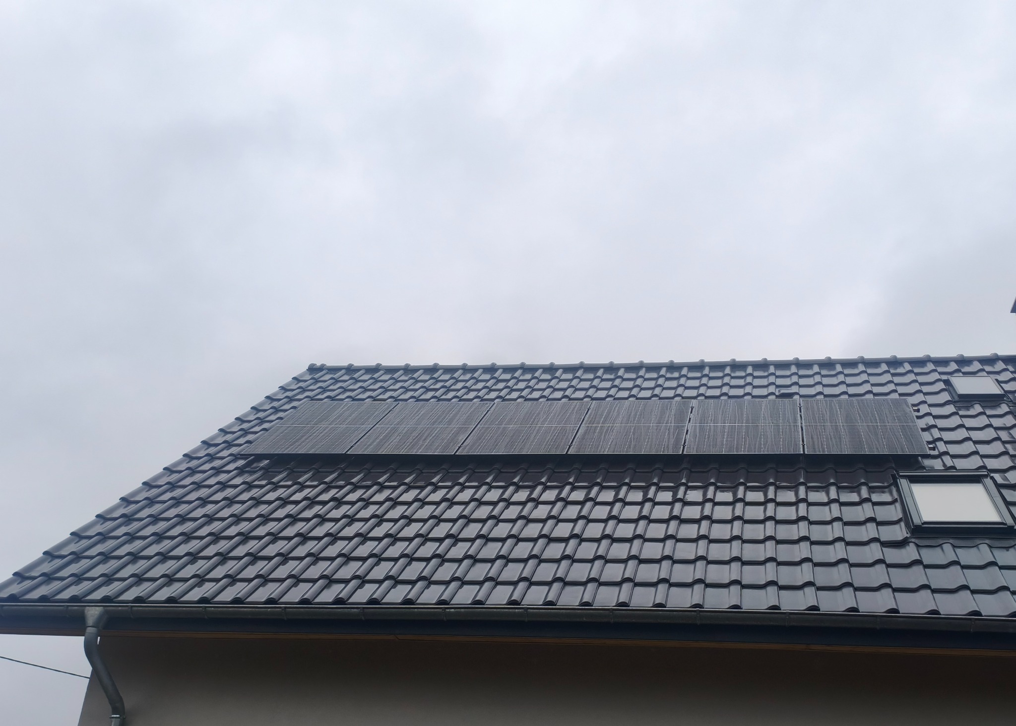 Panel słoneczny na dachu z ciemnej dachówki, widok z dołu pod kątem. Dach z oknami dachowymi, pochmurne niebo w tle. Widoczny fragment rynny i ściany budynku.