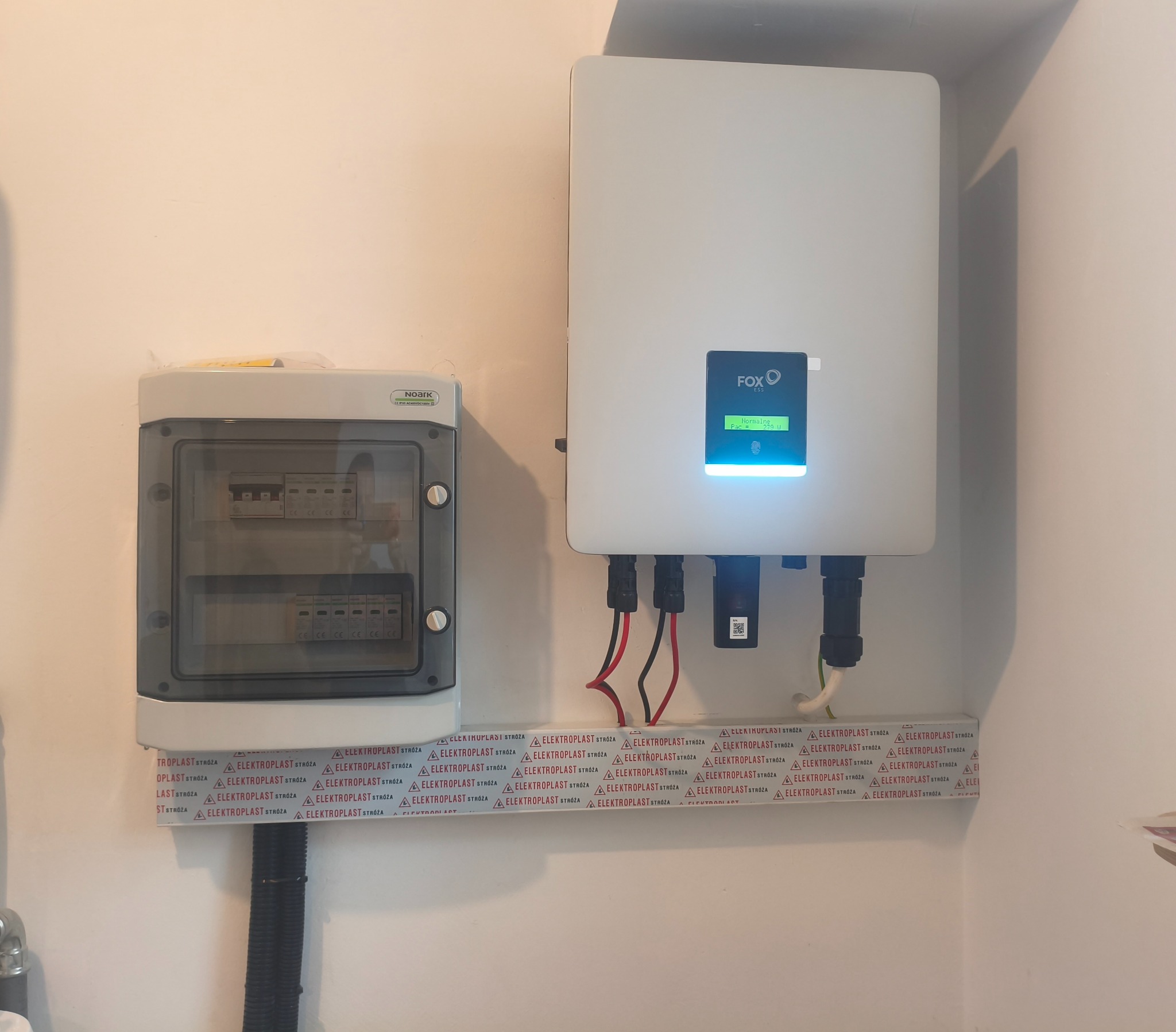 Biała skrzynka inwertera fotowoltaicznego Fox ESS z podłączonymi kablami, obok szara skrzynka z bezpiecznikami na ścianie, zabezpieczone listwą Elektroplast.