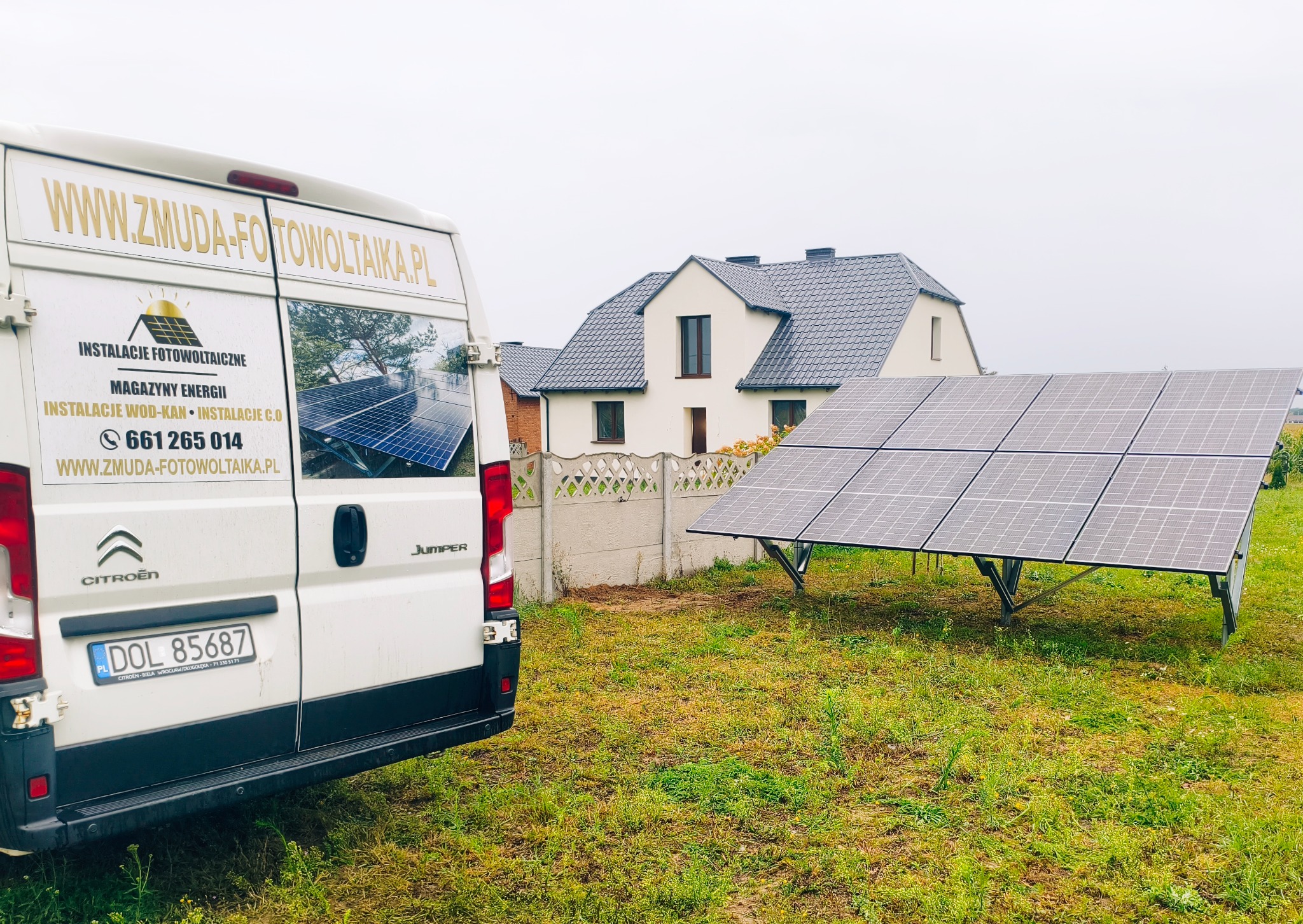 Instalacja paneli słonecznych na trawniku przed domem, obok zaparkowany biały bus z reklamą firmy fotowoltaicznej. Pochmurne niebo w tle.
