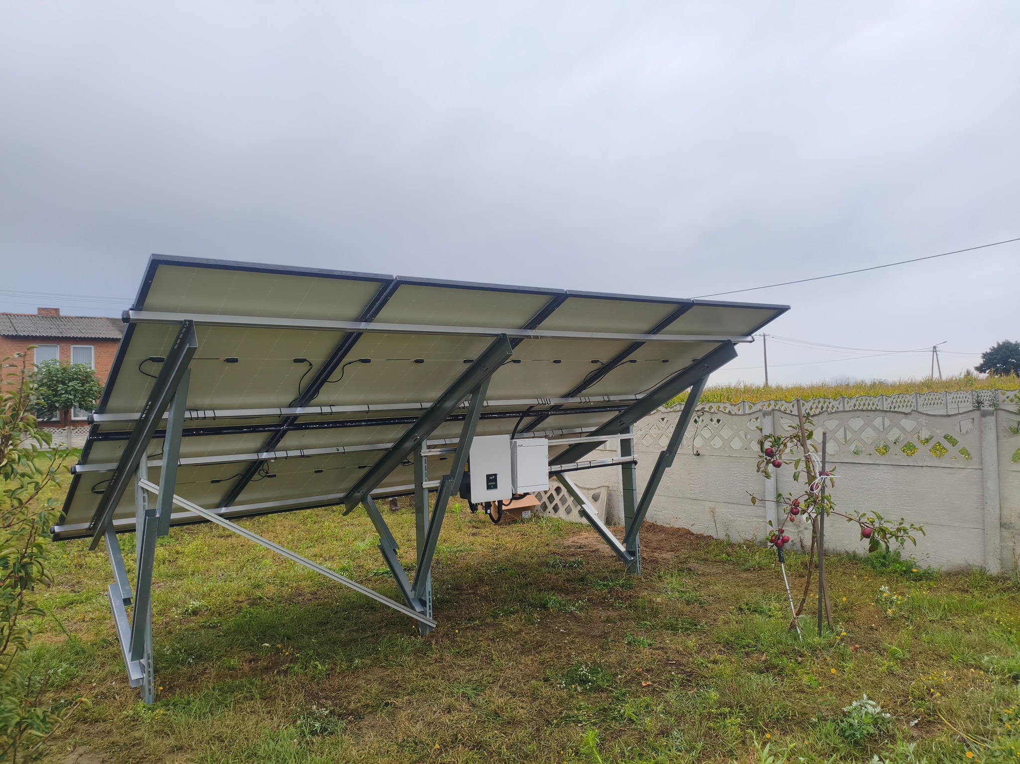 Instalacja fotowoltaiczna z widocznym inwerterem, zamontowana na trawniku obok ogrodzenia. Widok od spodu paneli, pochmurne niebo w tle.