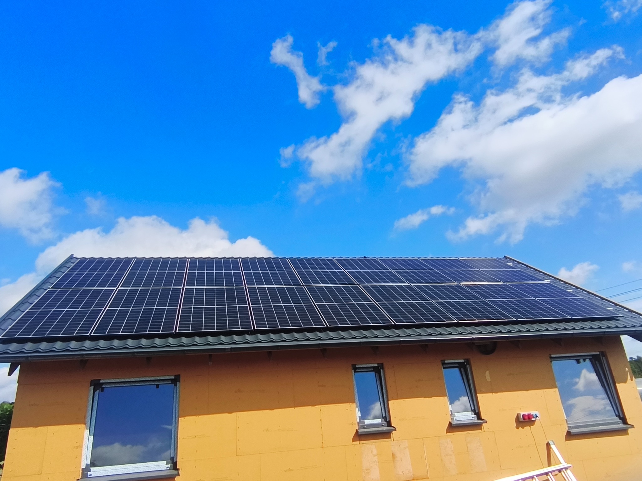 Dom w trakcie budowy z panelami słonecznymi na dachu, błękitne niebo z chmurami w tle. Widoczne okna z odbiciem nieba i drabina oparta o ścianę.