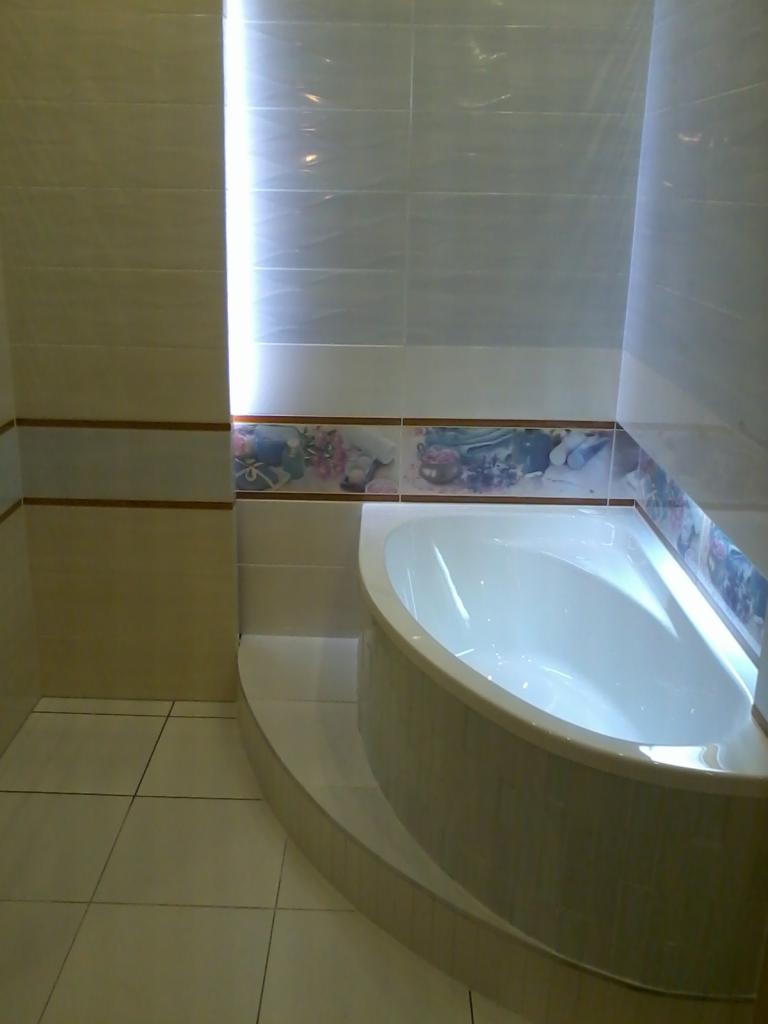 Wykończona łazienka z narożną wanną obudowaną płytkami w odcieniach beżu i bieli, dekoracyjny pas płytek z motywem kwiatowym na ścianie.