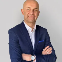 Uśmiechnięty mężczyzna w granatowej marynarce w kratę, białej koszuli i z zegarkiem na ręku, pozujący na jasnym tle.