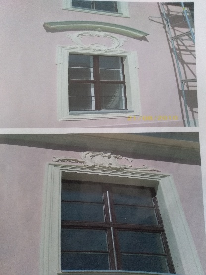 Dwa okna z ozdobnymi, jasnymi obramieniami i dekoracyjnymi elementami nad oknami na różowej elewacji budynku, widoczne rusztowanie z prawej strony.