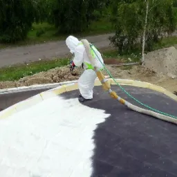 Pracownik w kombinezonie ochronnym aplikuje białą piankę poliuretanową na czarną membranę dachu, widoczny przew&oacute;d i otoczenie z ziemią i roślinnością.