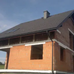 Dom w trakcie budowy: ceglana konstrukcja z zamontowanym dachem z dachówką, widoczne rynny, okno dachowe i komin. Budynek w stanie surowym otwartym.